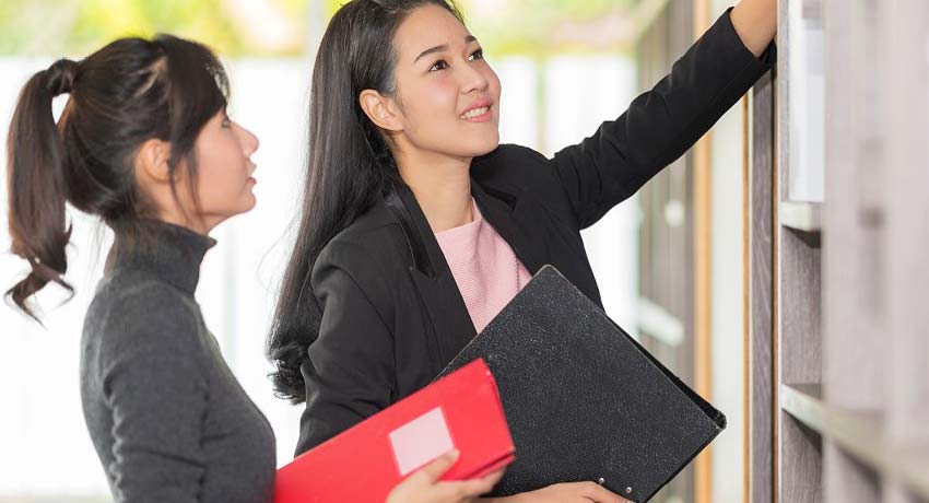 Two women in an office placing files on a shelf.