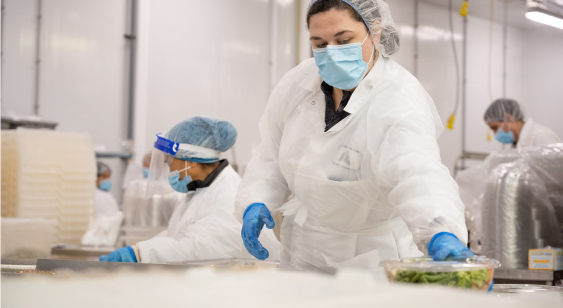 Workers in protective clothing and face masks handle food containers in a sterile processing environment.
