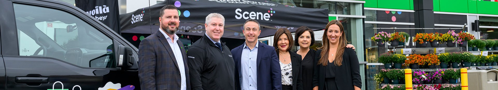 A group of six people stand together smiling in front of a black tent labeled "Scene+" and a background of a building and colorful flowers.
